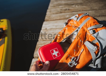 Close-up of orange backpack and red first aid kit placed on weathered wooden dock beside calm dark water, bright sunlight creating sharp shadows and highlighting straps, fabric texture, and zipper