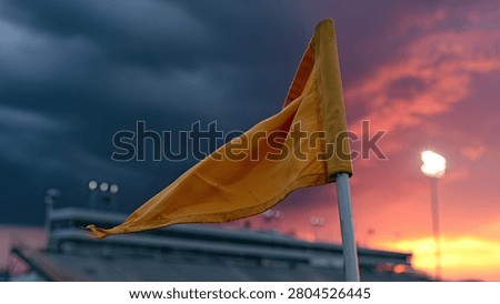 A close-up of an orange corner flag on a sports field, fluttering in the wind against a vibrant sunset sky with stadium lights in the background.