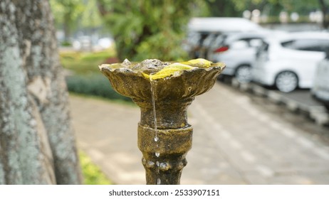 A close-up of an ornate, bronze fountain with a water droplet dripping from its spout. The fountain is adorned with intricate carvings and sits atop a stone pedestal. A few yellow petals float - Powered by Shutterstock