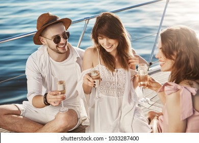 Close-up outdoor portrait of three friends talking and drinking champagne while sitting on board of boat and enjoying sunlight. Young couple and their friend discussing wedding ceremony