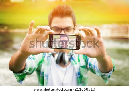 Closeup outdoors shot of young hipster man with headphones taking a selfie with smart phone by the river. Modern teenage guy taking a self portrait outdoors.