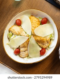 A close-up overhead view of a fresh Caesar salad featuring tender grilled chicken strips, vibrant cherry tomatoes, and delicate parmesan shavings
