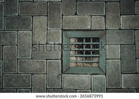 Close-up overhead view of grey interlocking concrete pavement with a square metal stormwater drain grate in the center. Textured urban ground, industrial floor design and public infrastructure