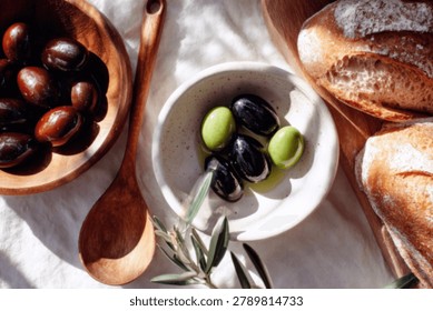 A close-up, overhead view of a rustic still life featuring a wooden bowl of Kalamata olives, a small white dish with green and black olives in oil, and crusty bread on a textured linen cloth.