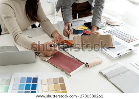 A close-up overhead view shows two pairs of hands belonging to Asian female designers — one in a beige sweater holding a pen — closely examining and selecting from a large spread of fabric swatches, material chips, and color sample cards on a white studio desk. A laptop and more color swatch books are visible at the edges, emphasizing a detail-focused, hands-on approach to interior design material selection.