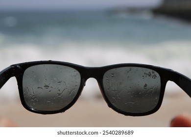 A close-up of a pair of black sunglasses with water droplets on the lenses. The sunglasses are in focus, while the background of a beach and ocean is blurry. - Powered by Shutterstock