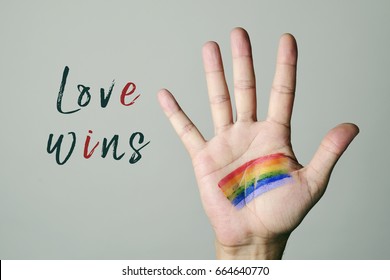 closeup of a the palm of a young caucasian man with a rainbow flag painted in it and the text love wins against an off-white background