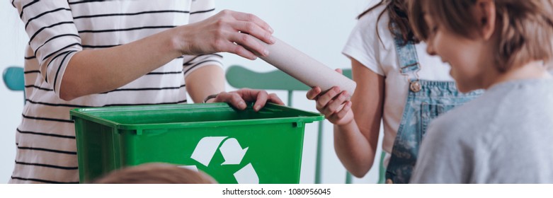 Close-up panorama of a school girl handing a paper tube to a teacher with a recycling bin during environment class for Earth Day - Powered by Shutterstock