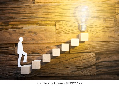 Closeup of paper businessman climbing block ladder leading towards illuminated light bulb on wood