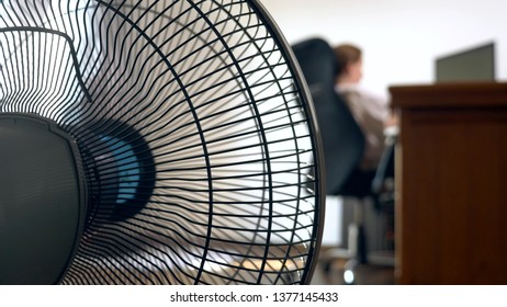 Close-up Of Part Of An Office Fan, On The Background Of A Blurred Writing Desk, At Which A Person Works At A Computer