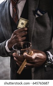 Close-up partial view of african american businessman holding glass of alcohol beverage and cigar while hiding dollar banknote in suit jacket pocket