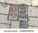 Close-up of pathway bricks with engraved letters and numbers, including "SGHS," "PP 1866," and "NO 8 MBP 1900," arranged on a stone-paved sidewalk.