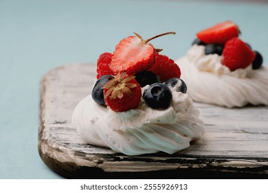 Close-up of pavlova desserts topped with fresh strawberries, blueberries, and raspberries, set on a rustic wooden board against a light teal background. Bright and appetizing focus on toppings. - Powered by Shutterstock