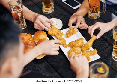 Close-up Of People Sitting At The Wooden Table Drinking Beer And Eating Fried Chicken Together In Cafe