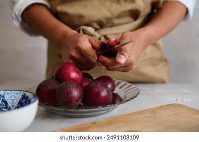 Close-up of a person in an apron slicing ripe plums on a wooden board with a glass bowl of plums nearby. Preparing fresh ingredients for a recipe in the kitchen. - Powered by Shutterstock