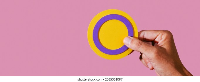 Closeup Of A Person With A Circular Intersex Flag In Their Hand On A Pink Background, In A Panoramic Format To Use As Web Banner Or Header