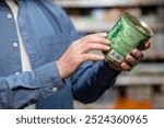 Close-up of person examining canned food label in supermarket. Focus on hands holding tin can, demonstrating careful consideration for product details in grocery shopping environment.