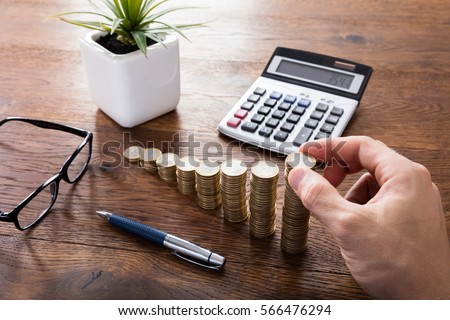 Close-up Of A Person Hand Calculating On Wooden Desk In Front Of Stacked Coins. Income Tax Raise Concept