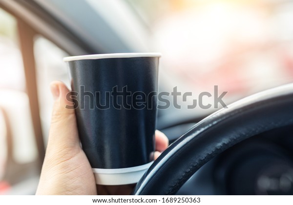 Close-up person hand holding black blank mockup\
paper cup woth hot coffee or tea takeaway drink driving in urban\
traffic jam city. Car dashboard and sunrise sunny window background\
early good morning