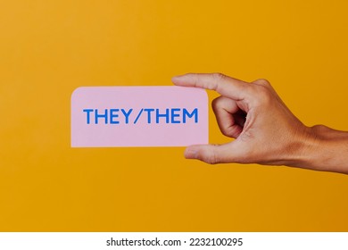 closeup of a person holding a pink sign with the pronouns they, them, against an orangish yellow background