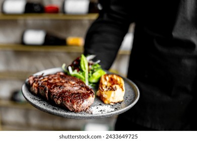 A close-up of a person holding a plate with a well-grilled steak, fresh greens, and a roasted half of garlic. The background is blurred, suggesting a kitchen or dining setting - Powered by Shutterstock