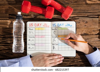 Close-up Of A Person Making Exercise Plan On Notebook Over Wooden Desk