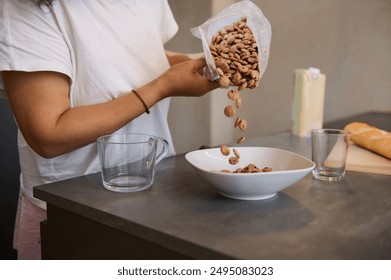 Close-up of a person pouring cereal into a bowl, with a jug and glasses on a kitchen countertop. Home breakfast preparation scene in a cozy kitchen. - Powered by Shutterstock