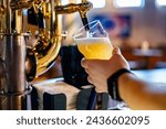 Close-up of a person pouring fresh, frothy beer from a tap into a glass in a cozy bar setting, with blurred wooden furniture in the background