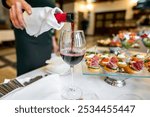 A close-up of a person pouring red wine into a glass, with appetizers in the background. image captures the elegance of fine dining, highlighting the rich color of the wine and inviting atmosphere.