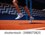 Close-up of a person running on a red track in a stadium. The runner is wearing white and blue running shoes and black shorts. The background features a blue and white geometric pattern on the stadium