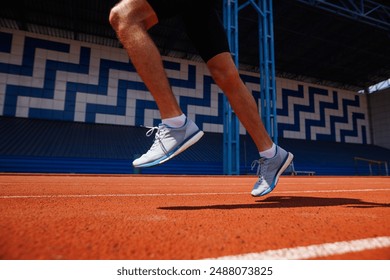 Close-up of a person running on a red track in a stadium. The runner is wearing white and blue running shoes and black shorts. The background features a blue and white geometric pattern on the stadium - Powered by Shutterstock