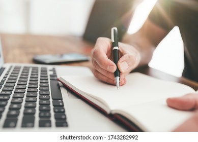 close-up of person taking notes with pen on paper in front of laptop computer