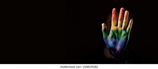 closeup of a person with their hand patterned with the rainbow pride flag in front of their face, on a black background, in a panoramic format to use as web banner or header