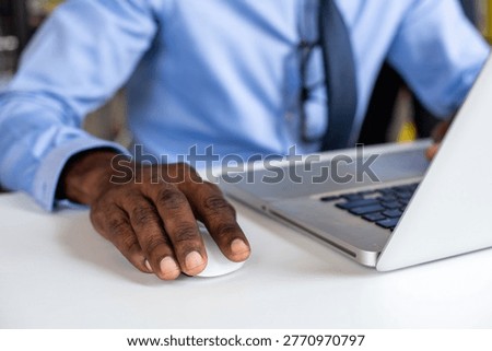 Close-up of a person using a computer mouse with a laptop, wearing a blue shirt and tie, at a white desk in an office setting.