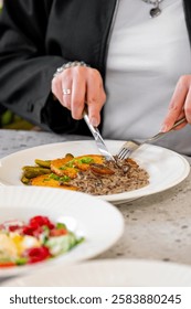 A close-up of a person using a knife and fork to cut into a plate of delicious food, including meat, vegetables, and a creamy sauce, set against a soft-focus background.