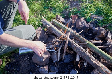 Close-up person using small butane torch to light campfire. Fire pit arranged with stones and stack of logs and twigs, situated on grassy field. - Powered by Shutterstock