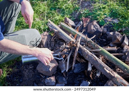 Close-up person using small butane torch to light campfire. Fire pit arranged with stones and stack of logs and twigs, situated on grassy field.
