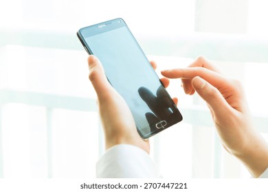 Close-up of a person using a smartphone while standing by a bright window in a modern setting