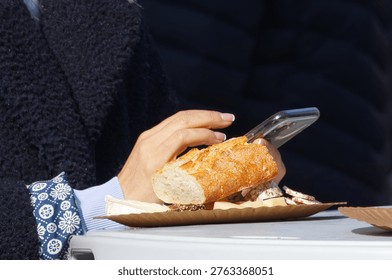 Close-up of a person using a smartphone while enjoying fresh baguette and cheese at the Naplavka market. Perfect for modern lifestyle, outdoor dining, and street food concepts.A detailed close-up shot capturing a modern outdoor dining moment at the Naplavka market in Prague. In the foreground, a paper plate rests on a white table, holding a crusty piece of French baguette, assorted cheeses including camembert, and sliced cured meat. Behind the food, a woman's hands with manicured nails are seen holding and actively typing on a mobile phone. She is wearing a cozy dark textured fleece jacket over a blue patterned shirt. The scene highlights a realistic blend of culinary enjoyment and digital connectivity in a relaxed urban setting.