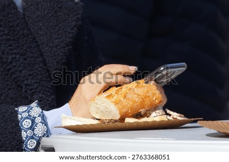 Close-up of a person using a smartphone while enjoying fresh baguette and cheese at the Naplavka market. Perfect for modern lifestyle, outdoor dining, and street food concepts.A detailed close-up shot capturing a modern outdoor dining moment at the Naplavka market in Prague. In the foreground, a paper plate rests on a white table, holding a crusty piece of French baguette, assorted cheeses including camembert, and sliced cured meat. Behind the food, a woman's hands with manicured nails are seen holding and actively typing on a mobile phone. She is wearing a cozy dark textured fleece jacket over a blue patterned shirt. The scene highlights a realistic blend of culinary enjoyment and digital connectivity in a relaxed urban setting.