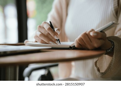 Close-up of a person writing notes in a notebook beside a laptop in a modern café, representing study, planning, and productivity.