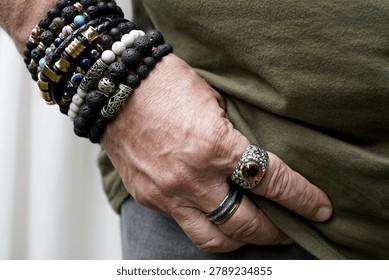 A  close-up of a person's hand adorned with multiple bracelets and rings. The bracelets, made of black lava beads, stones, and metallic elements, are intricately layered. Two rings are worn—a blue band on the index finger and a large ornate ring with an amber-colored gemstone on the middle finger. The hand is positioned casually in the pocket of a grey fabric