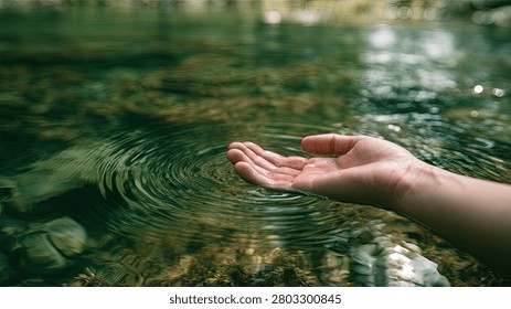 A close-up of a person's hand gently touching the surface of a clear, shallow forest stream, creating soft ripples.