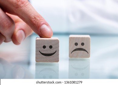 Close-up Of Person's Hand Holding Wooden Block With Happy And Unhappy Face On Reflective Desk