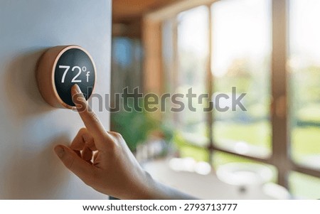 Close-up of a person's hand setting a smart thermostat to 72 degrees Fahrenheit, symbolizing energy efficiency, smart home technology, and lifestyle comfort in a bright, modern living space. The sunlight streaming through large windows creates a warm and inviting atmosphere, emphasizing innovation, sustainability, and everyday convenience in home automation.
