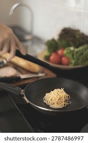 A close-up of a person's hand using black tongs to place plain, cooked spaghetti into a frying pan on a stovetop. Fresh vegetables and bread are blurred in the kitchen background.