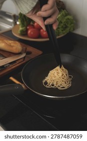 A close-up of a person's hand using black tongs to place plain, cooked spaghetti into a frying pan on a stovetop. Fresh vegetables and bread are blurred in the kitchen background.