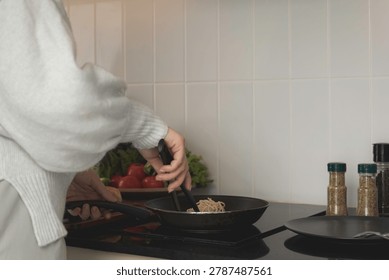 A close-up of a person's hand using black tongs to place plain, cooked spaghetti into a frying pan on a stovetop. Fresh vegetables and bread are blurred in the kitchen background.