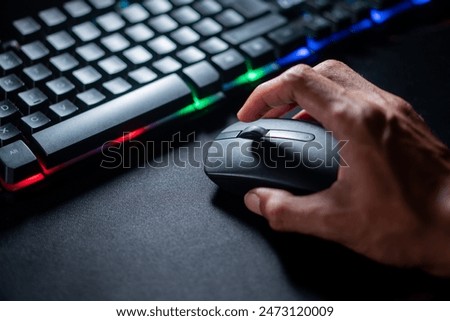 Close-up of a person's hand using a computer mouse next to a keyboard with RGB lighting on a dark surface.