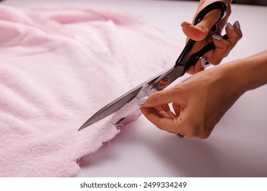A close-up of a person's hand using large scissors to cut a piece of pink fabric on a white surface. The focus is on the scissors and the fabric, showcasing a crafting or sewing activity.